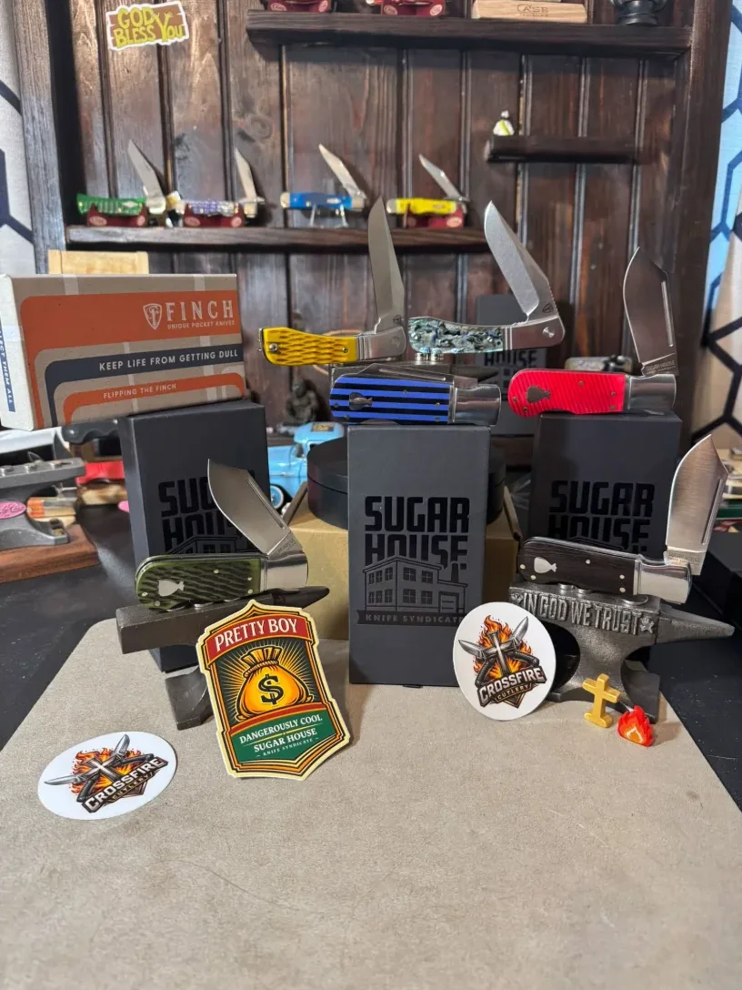 Crossfire Cutlery's own display shelf: traditional pocket knives on a dark wood shelf, a "God Bless You" sign, boxed Sugar House knives, Crossfire stickers, and a small anvil stamped "In God We Trust"