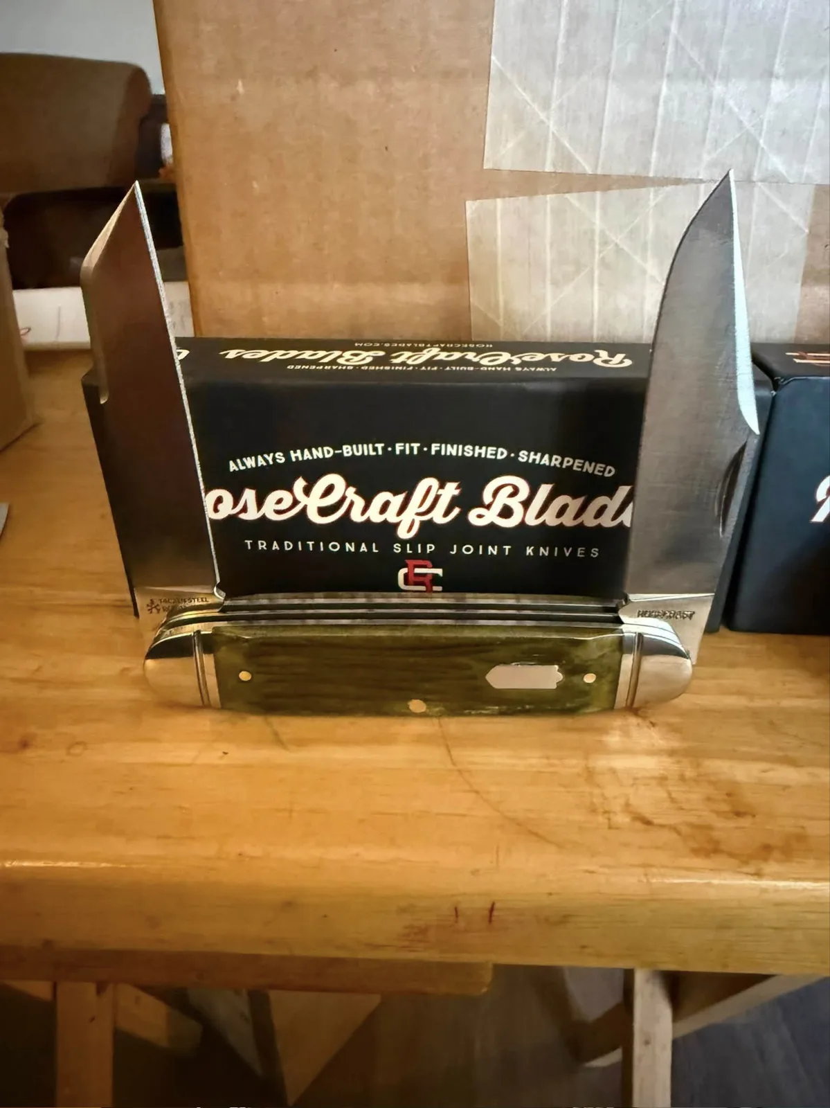 A RoseCraft Blades trapper with green bone handles, both blades open, in front of its RoseCraft box on a wood workbench