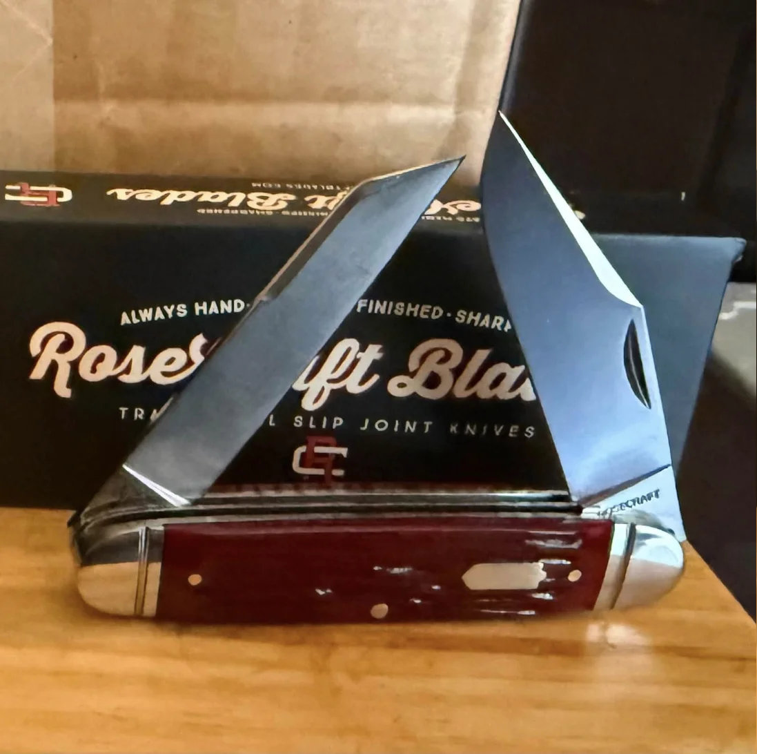 A real RoseCraft Blades trapper pocket knife, open and photographed on a wood table in front of its own packaging — Crossfire Cutlery's own inventory